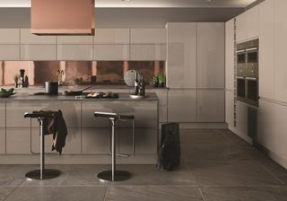 Roma Cashmere High Gloss kitchen with a kitchen island, bar stools and copper backplash, lighting and stone slab floor tiles by Optiplan Kitchens