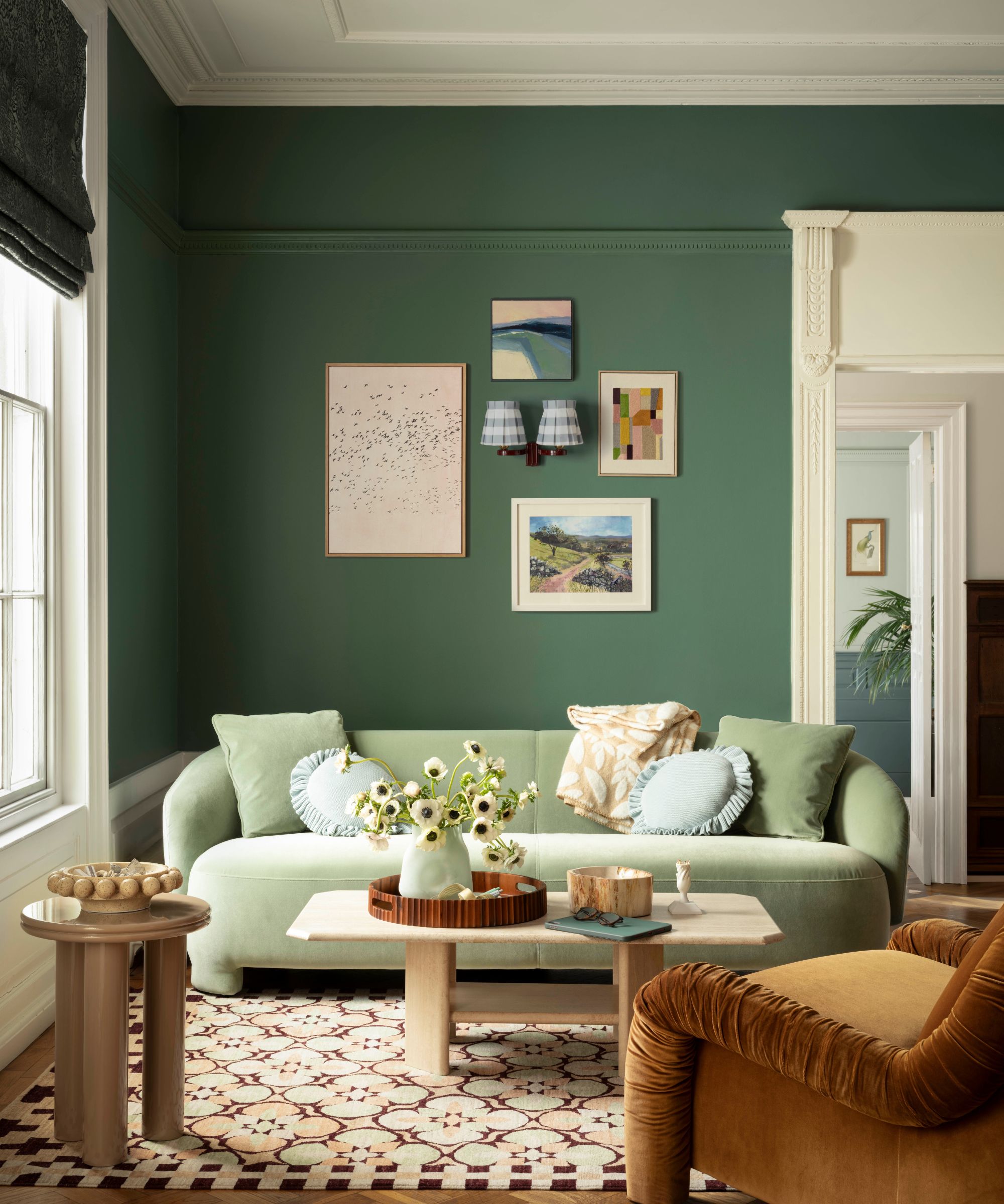 A living room is painted in Webster Green, with a pale green velvet sofa, a patterned rug, a brown velvet armchair, and a gallery wall of prints.