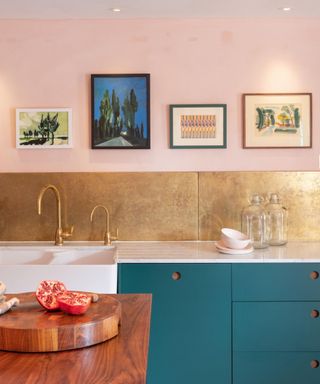 A kitchen with light pink walls with four wall art prints, a gold backsplash underneath it, teal cabinets with a white built-in sink, and a dark wooden kitchen island with a chopping board with tomatoes on it