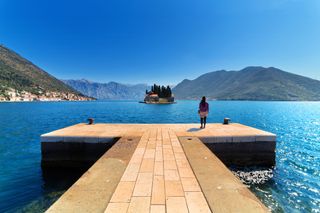 Lone female tourist standing on dock at Our Lady of the Rocks (Gospa od Skrpjela) man-made island, looking across water to Sveti Dorde Island with coastal town of Perast in distance on Bay of Kotor, Montenegro