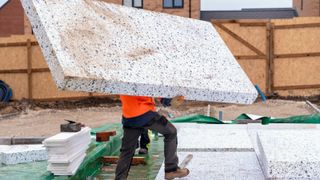 A man in a red top carrying a huge slab of polystyrene insulation across a building site