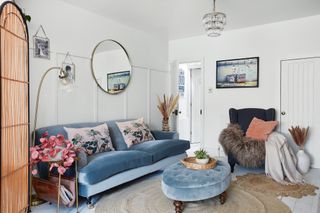 Living room with white panelled walls, blue velvet sofa and footstool, round wall mirror, brass floor lamp and jute rug