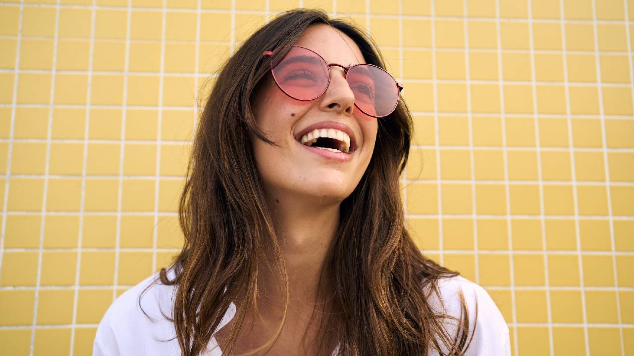 A woman smiling while wearing a pair of round, red-tinted sunglasses.