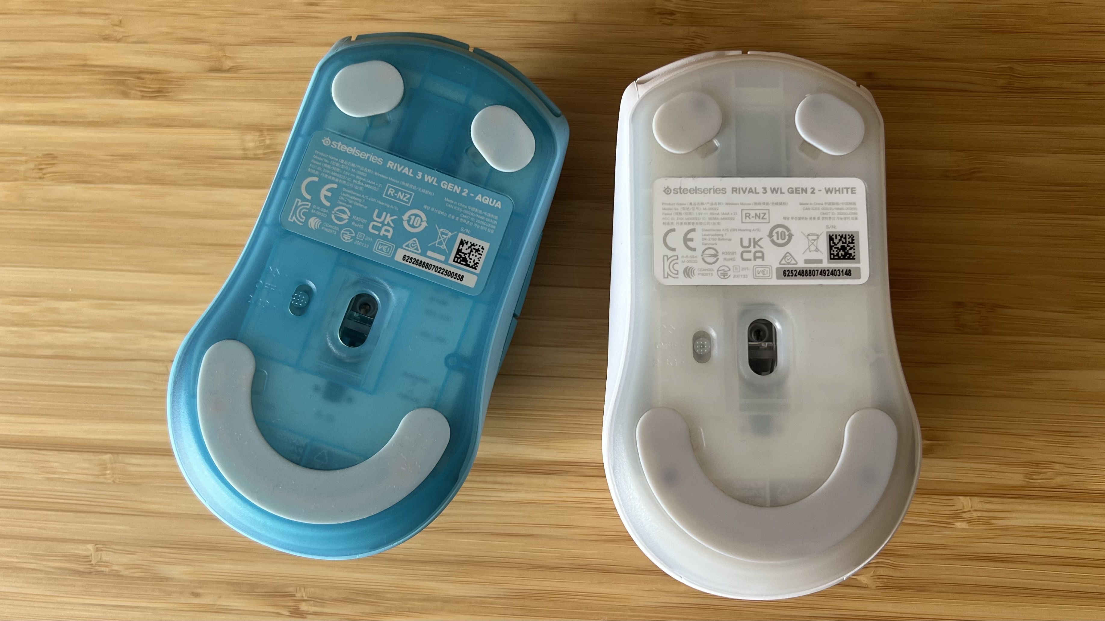 Underside of a blue SteelSeries Rival 3 Wireless Gen 2 gaming mouse next to a white model, the translucent effect is more pronounced on the blue version