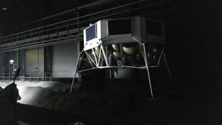 An image of a gray metal hexagonal lunar lander with four thin metal feet standing in a dimly lit warehouse with gray rocks on the floor