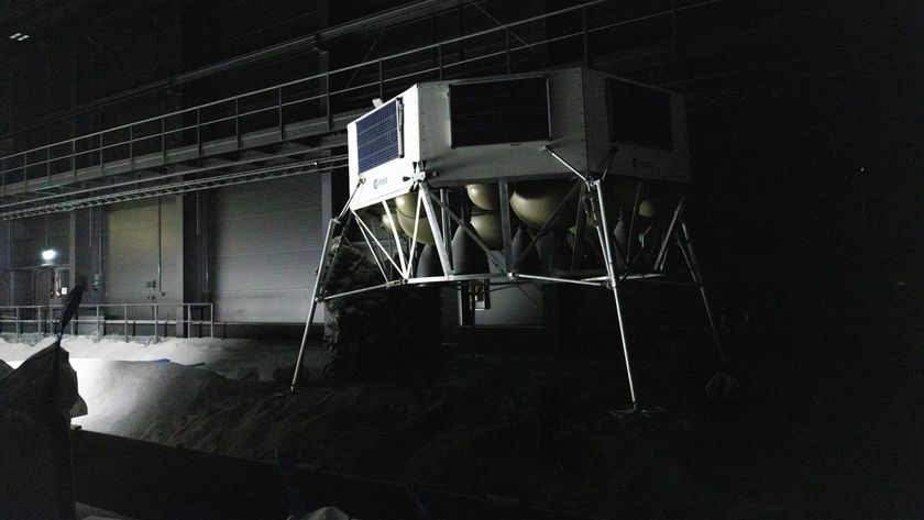 An image of a gray metal hexagonal lunar lander with four thin metal feet standing in a dimly lit warehouse with gray rocks on the floor