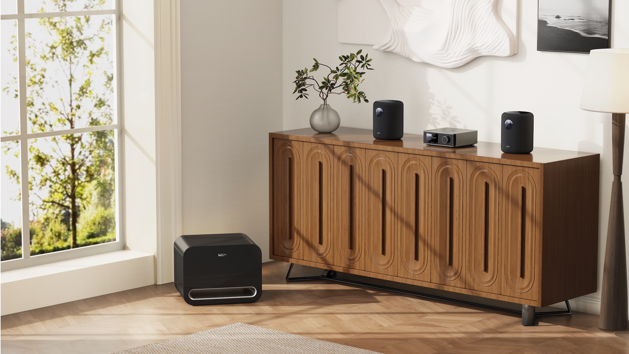 Two WiiM Sound smart speakers on a wooden sideboard in a brightly lit room. The Wiim Sub Pro is on the floor beside the sideboard and the WiiM Amp Ultra is between the speakers. 