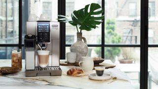 The De'Longhi Dinamica Plus bean-to-cup coffee machine on a kitchen countertop surrounded by coffees and pastries