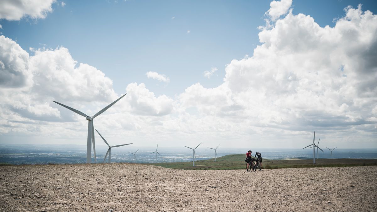 Two cyclists stand in a wind farm overlooking Manchester