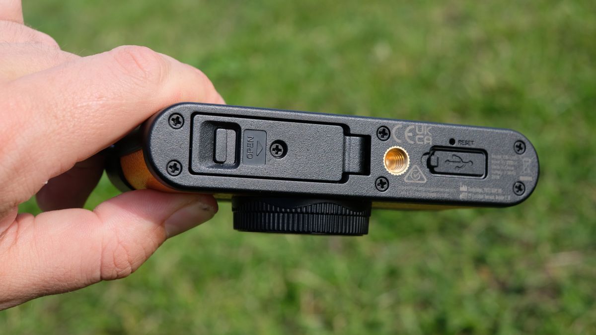Photograph of man's hand holding Camp Snap 2 digital camera, in yellow colorway, bottom plate showing battery and card slot door screwed in place, as well as tripod thread