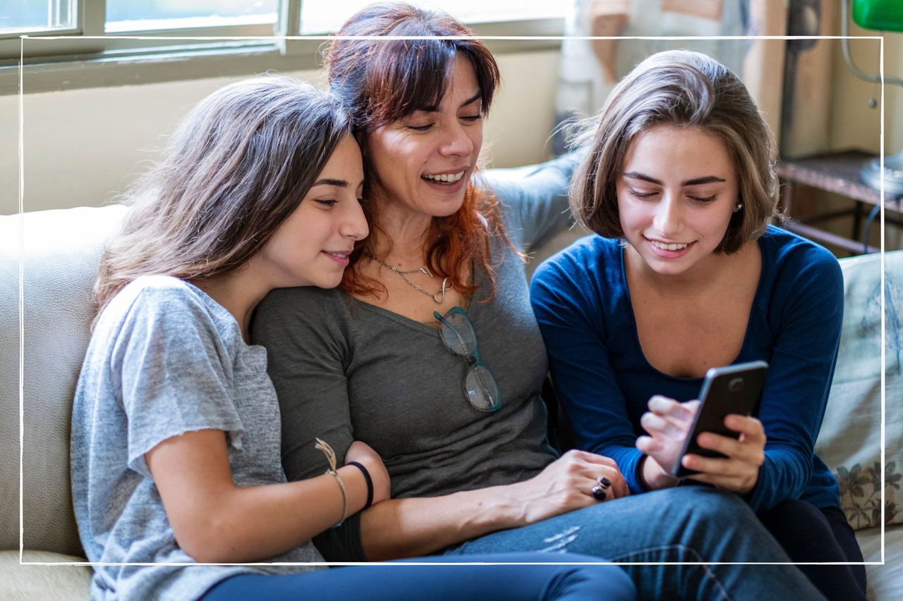 Mother sitting on sofa with teen daughters, they are all looking at a phone, doing some online shopping