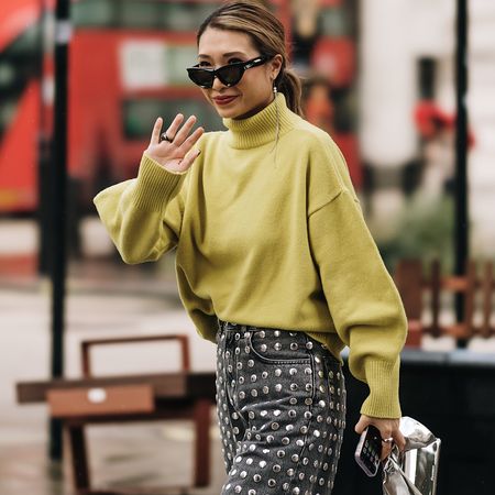 fashion week attendee wears sunglasses, green sweater, and studded jeans, waving 