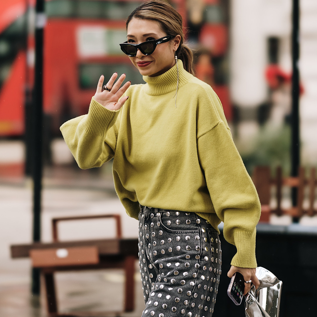 fashion week attendee wears sunglasses, green sweater, and studded jeans, waving 
