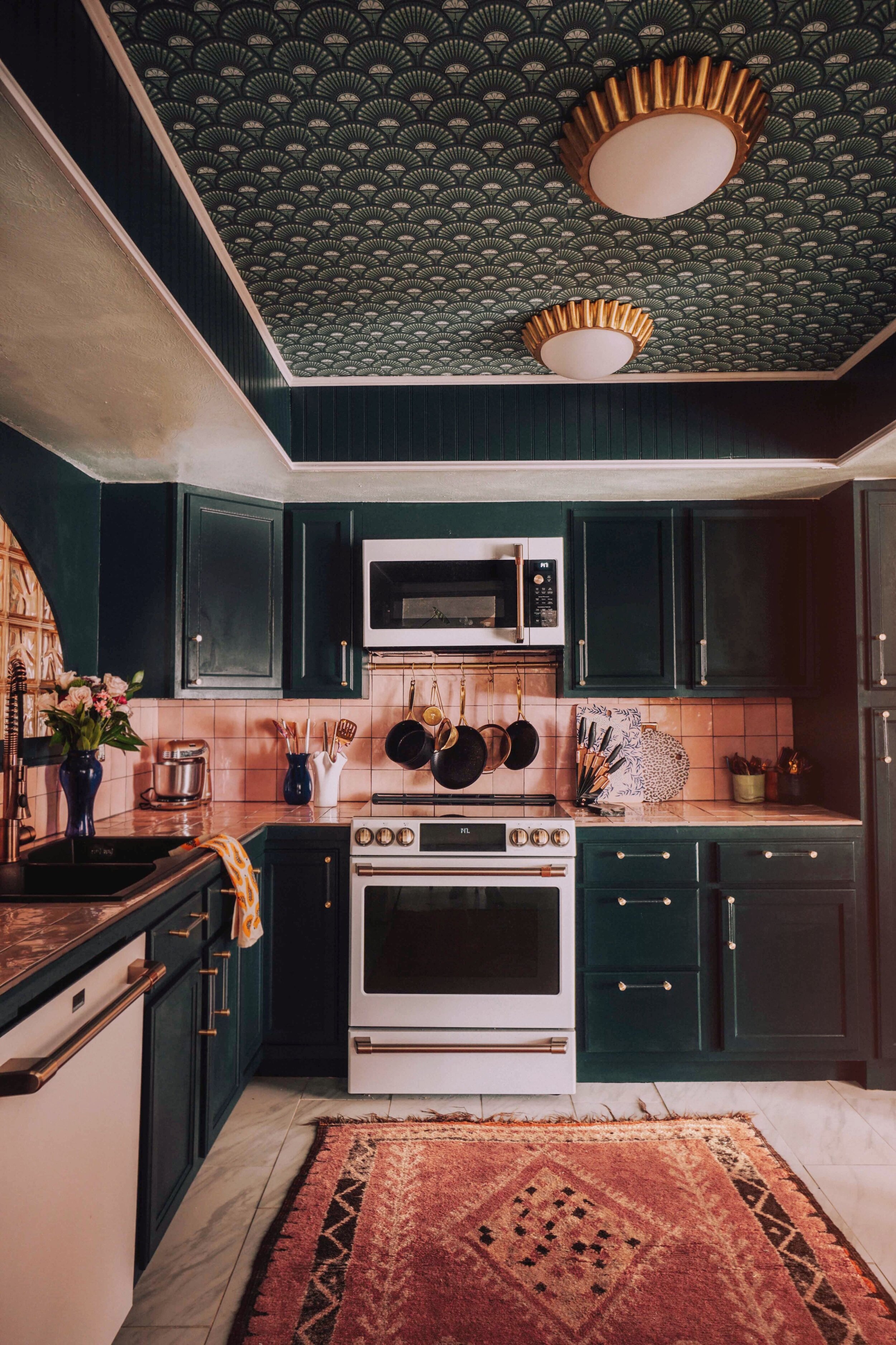 an art deco kitchen with dark cabinets, pink backsplash, and ceiling wallpapered in art deco print wallpaper with brass flush lighting