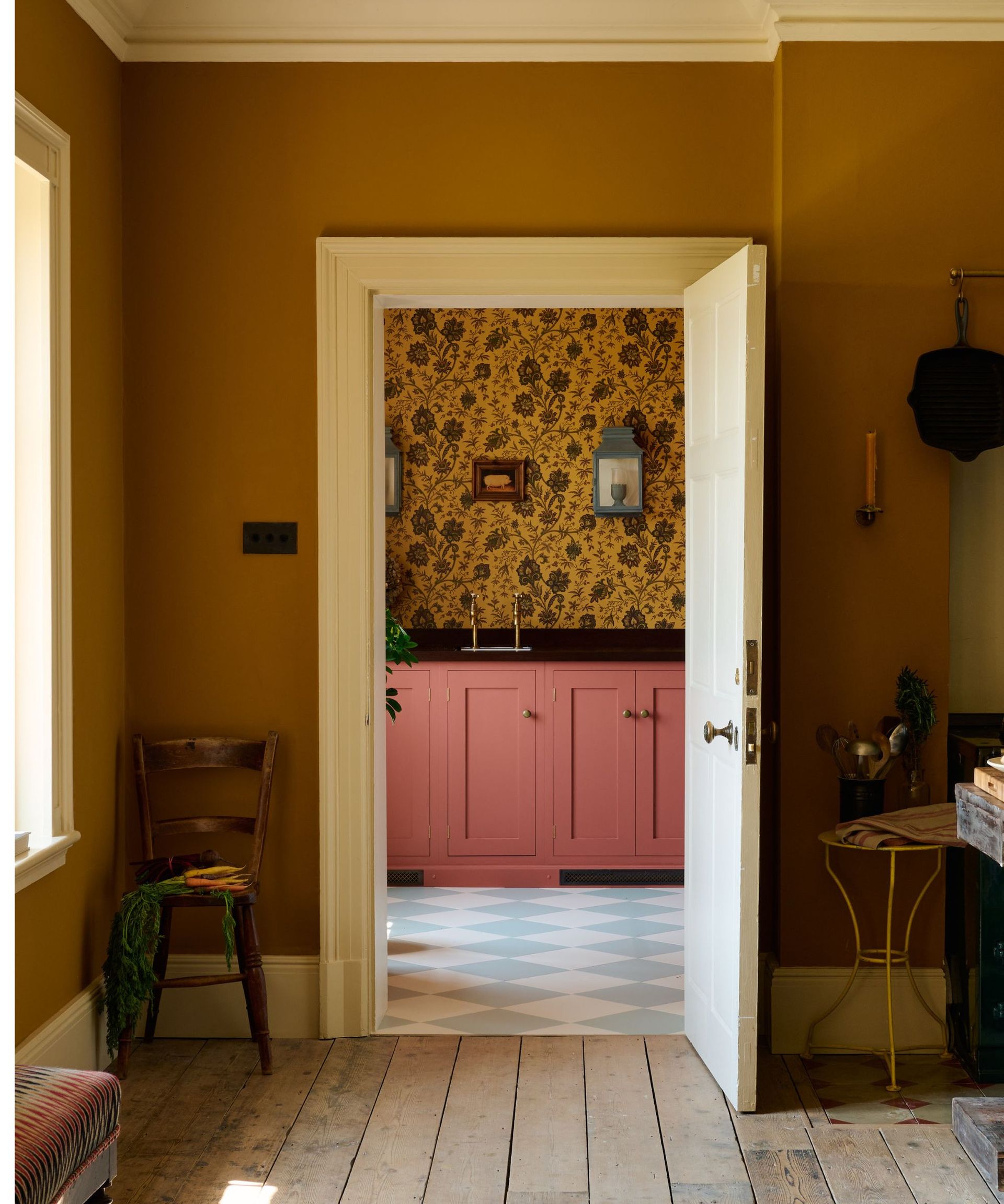 Dark mustard painted hallway with original wooden floors, showing a peek of pink kitchen cabinets and dark yellow wallpaper beyond. The kitchen floor has a white and light blue diamond pattern
