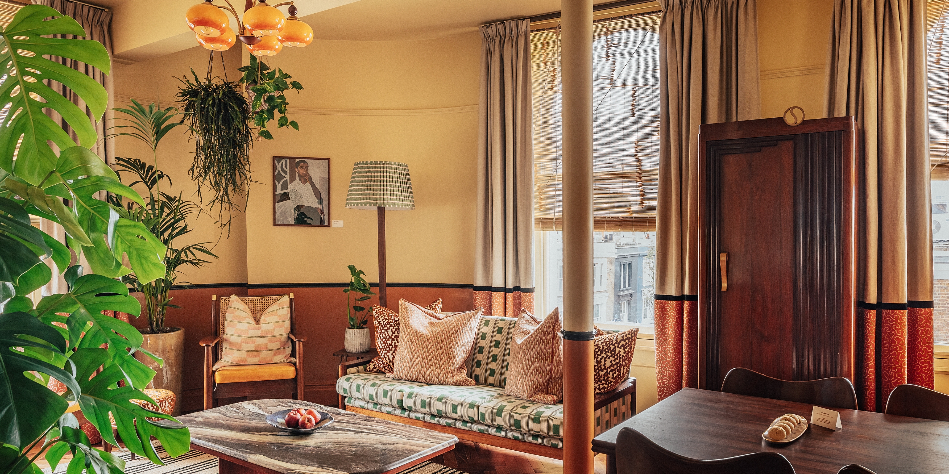 An ochre and wood-tinted hotel room features vibrant, upholstered armchairs and sofas with summery motifs, marble-topped coffee tables, and orante lamps, curtains, and artwork, along with thriving plants.