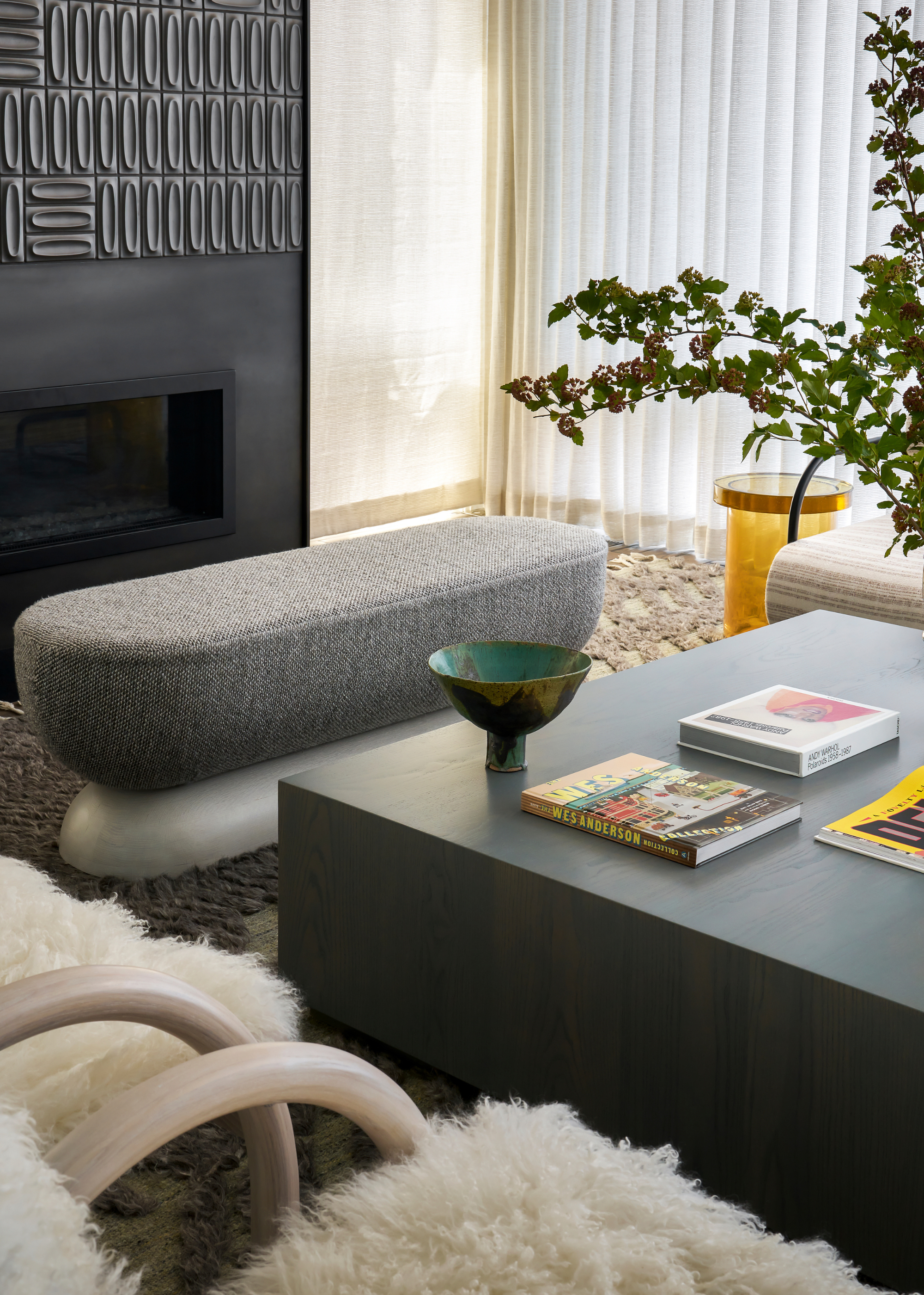 A living room with a black fireplace and accent wall, a textured area rug, a gray bench, a wood coffee table with books and vases, a yellow side table, and a pair of fuzzy armchairs