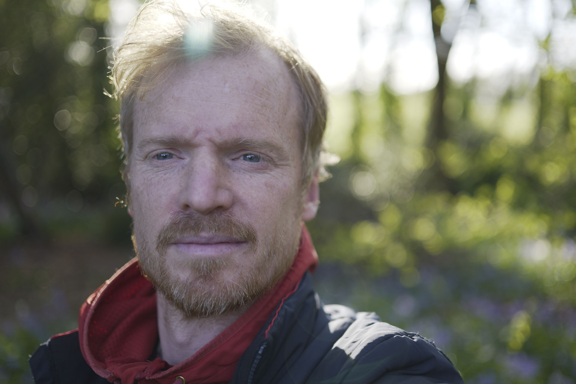 Man in an orange hoodie with strong sunlight behind him and dappled light through trees