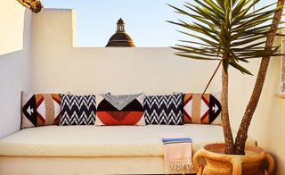 An outdoor sitting area with a long sofa, patterned cushions and a potted plant.