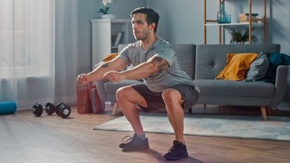 Man learning how to do squats in his living room; behind him we see a set of dumbbells, a yoga mat and a modern sofa