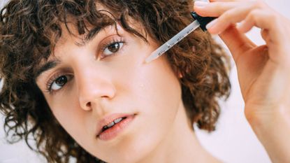 best face serum - woman with shorted curly hair looking into the camera with a pipette of serum next to her face