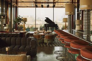 A 1970s-inspired, rooftop salon features sculptural upholstered seating including sofas, shearling armchairs, and bordeaux and chrome stools, as well as statement lighting, plants, and a grand piano.