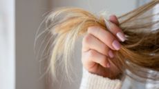 A woman's hand holding the ends of her blonde hair , with pink nails and a cream jumper