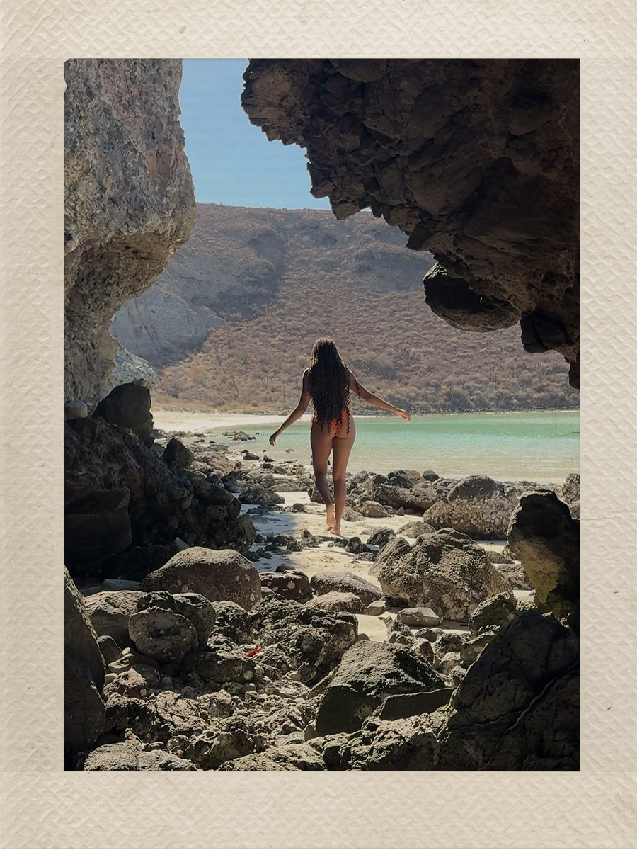 Claire Most steps across a rocky shoreline in a orange bikini