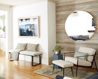 Living room with exposed wooden paneling and large round mirror mounted on wall, white painted walls with beige painted skirting board, cream and black armchair and bench with seat cushions, light wood flooring and rug, blue side table and wall art