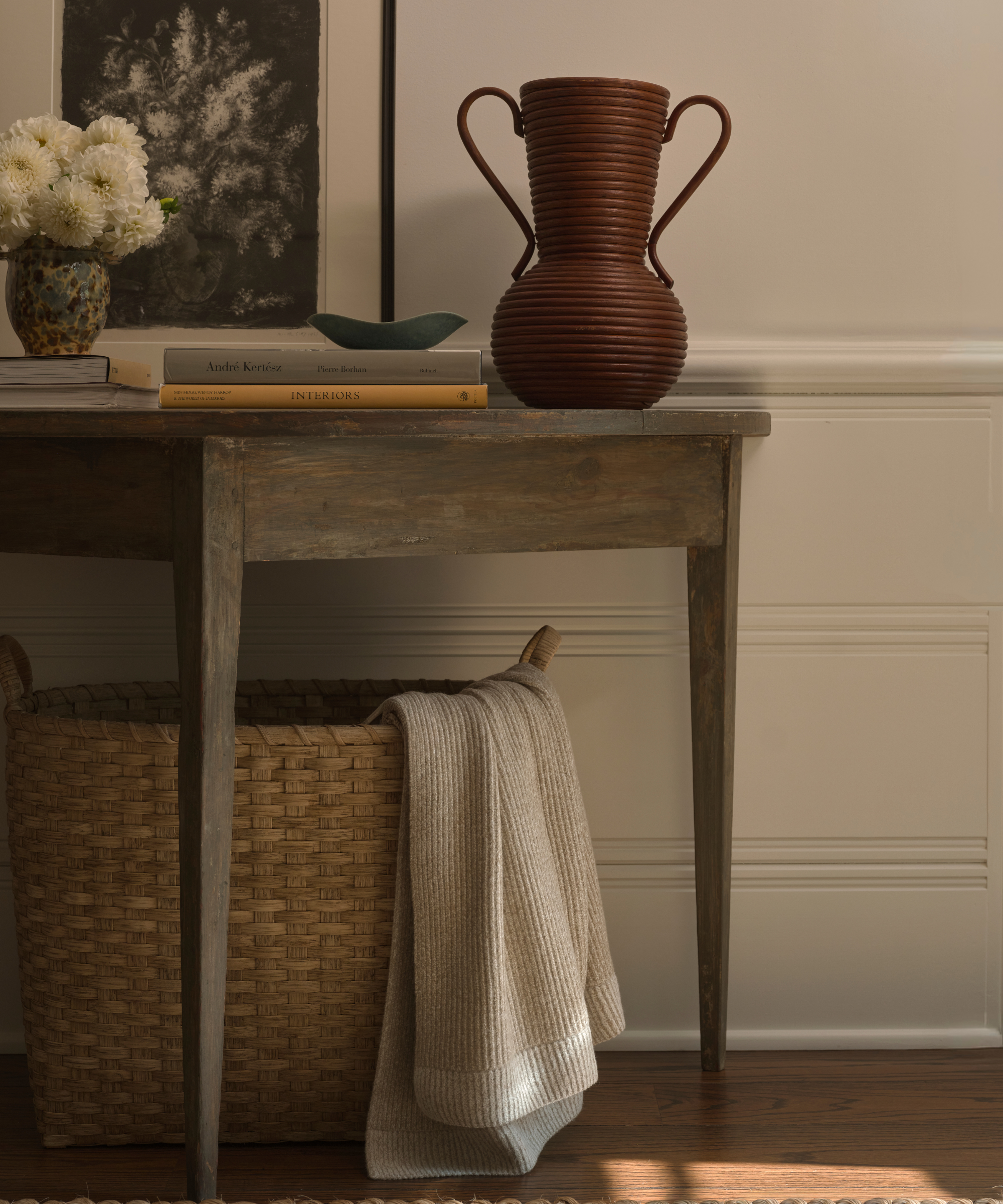 Cozy, neutral entryway vignette featuring a weathered wood entry table, larger woven basket with oatmeal woven throw, rattan vase, coffee table books, and floral artwork.