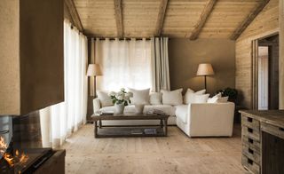 Interior view of a living area at San Luis featuring brown and wood panel walls, wooden beams, large windows with white curtains, a fireplace, a white corner sofa, a coffee table with white vase and flowers and two floor lamps