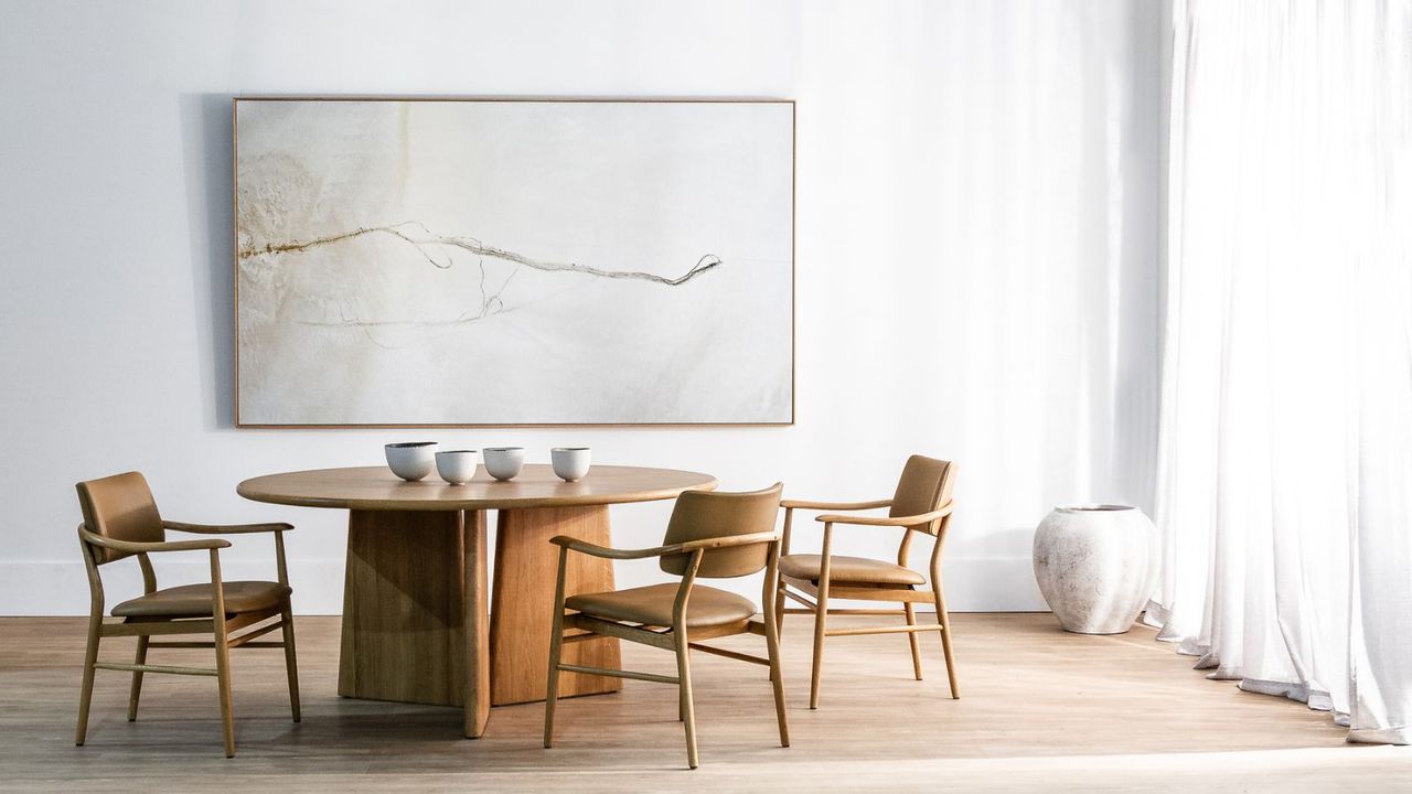 A natural teak dining table with three wide legs in the center of a white room with three teak chairs around it