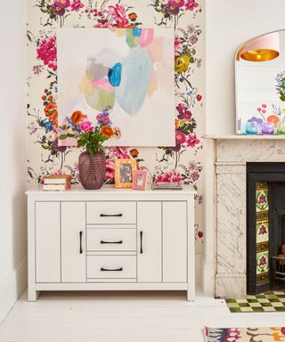 A white dining area with a colorful floral wall with a pastel wall art print, a white sideboard with a vase of pink flowers and photo frames, and a fireplace to the right with a gold arched mirror above it
