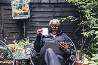 Man holding mug looking at digital tablet while sitting on chair in backyard
