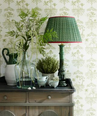 A green patterned table lamp on a console table with green and white patterned wallpaper.