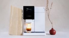 Image of a white, high-tech coffee machine brewing an espresso shot with a piece of beige stone and a small red flower vase on either side of it.