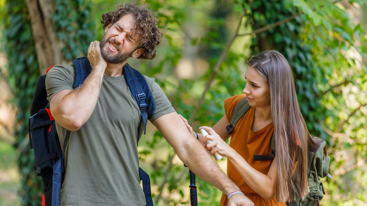 Man scratching neck in the woods while woman applies bug spray to his arm