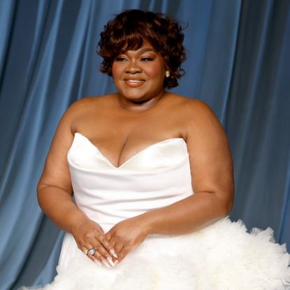 a black woman with short brown hair poses in a white strapless gown in front of a blue backdrop