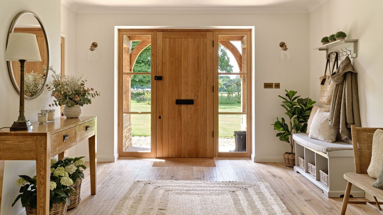 A large naturally lit entryway with a wooden front door framed by large windows. Light wooden plank flooring with a neutral boho rug. A console table on the left and coat storage on the right. 