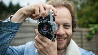 Man wearing blue denim jacket holding Nikon FM and looking through the viewfinder 