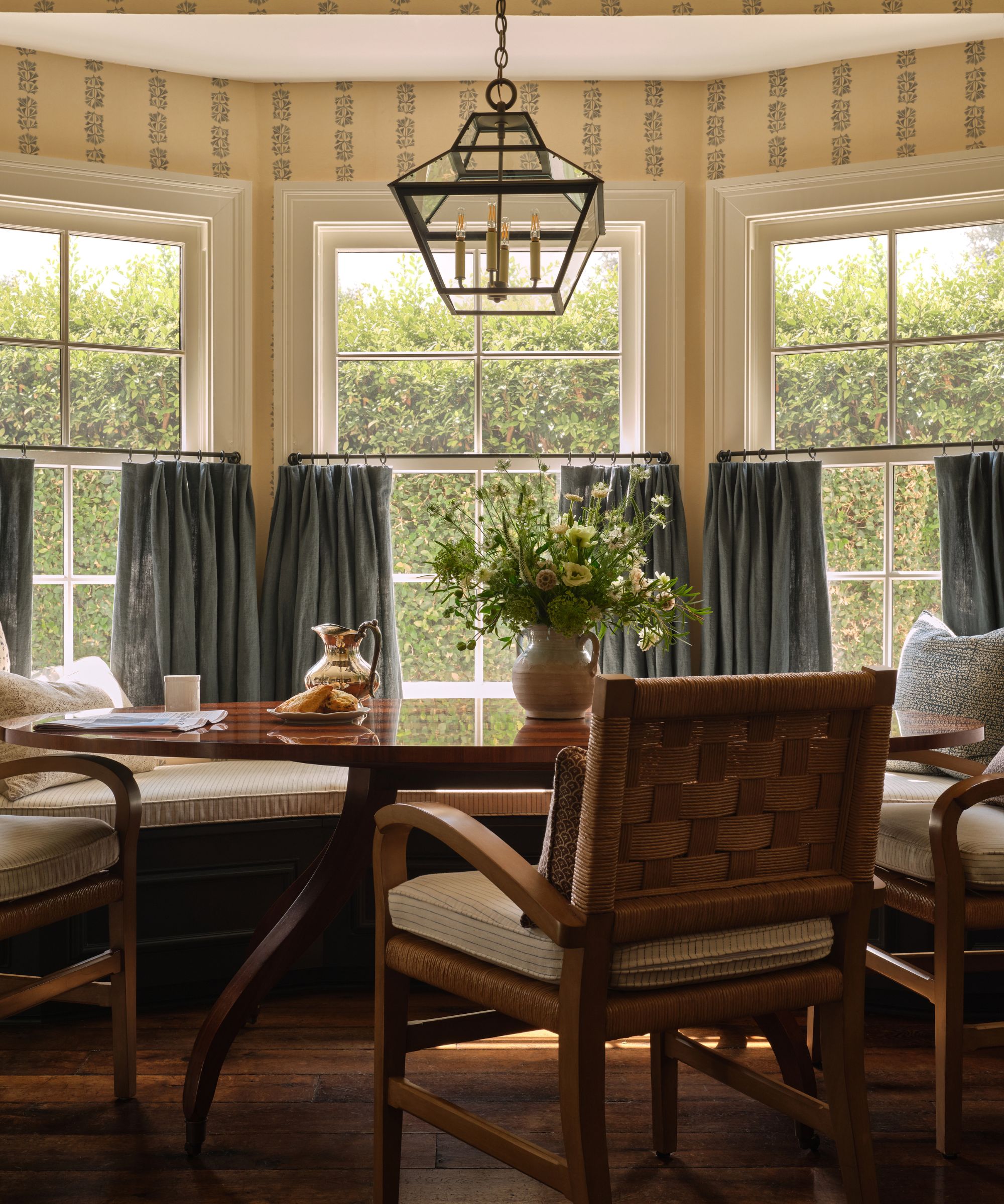 A breakfast nook with pale yellow wallpaper, bay windows with blue cafe curtains and a large dining table and chairs.