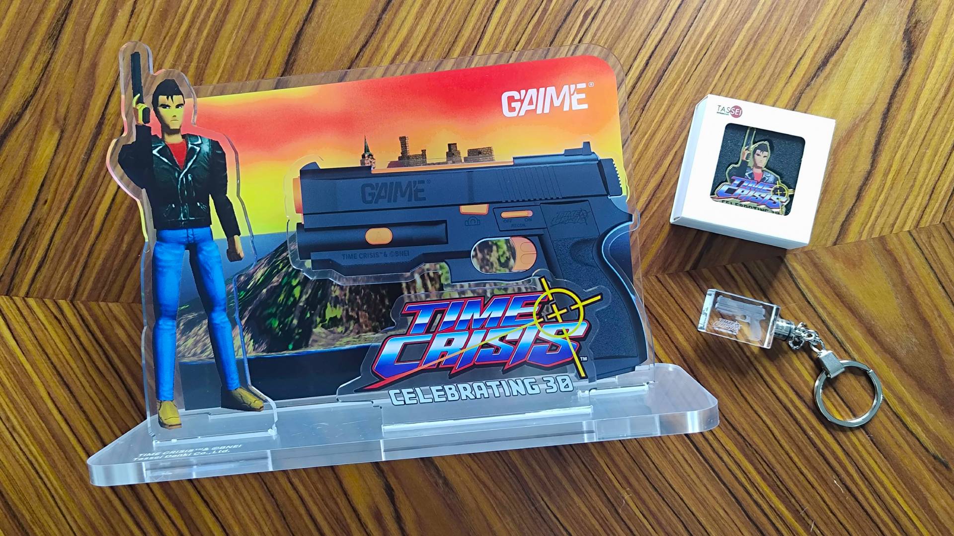 G&#039;AIM&#039;E Time Crisis console Ultimate Bundle extras featuring diorama, pin badge, and key chain on woodgrain table.
