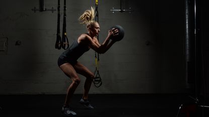 Best HIIT workout gear: pictured here, an sporty woman smashing a slam ball to the ground in a dark gym environment