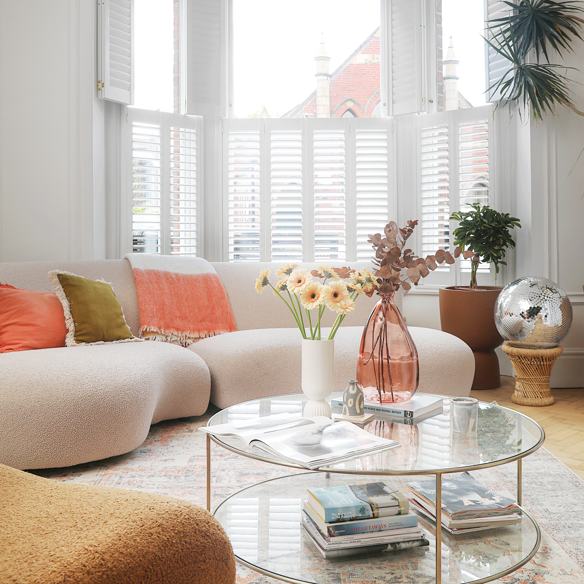 a living room with curved sofas and a round glass coffee table, window shutters and a glitter ball