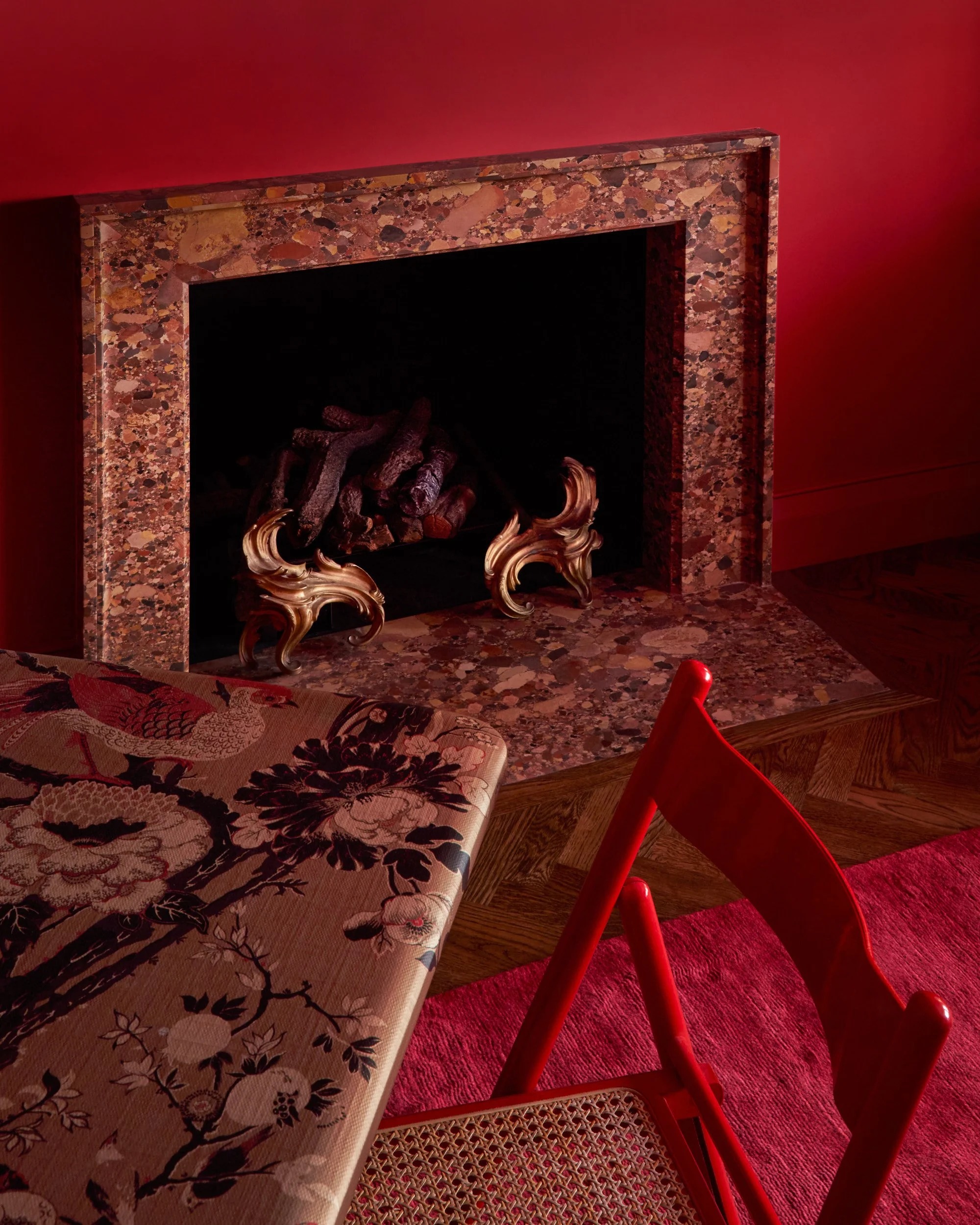 Image of a red room with a red granite fire place, a red rug, a floral game table, and a red and wicker dining chair.