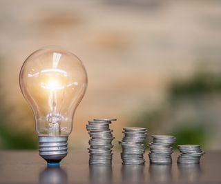 lightbulb stood upright on table with 4 piles of silver coins next to it, with each pile getting smaller