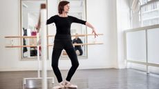 A woman stands in a ballet studio holding a bar with one hand and gently bending her knees.