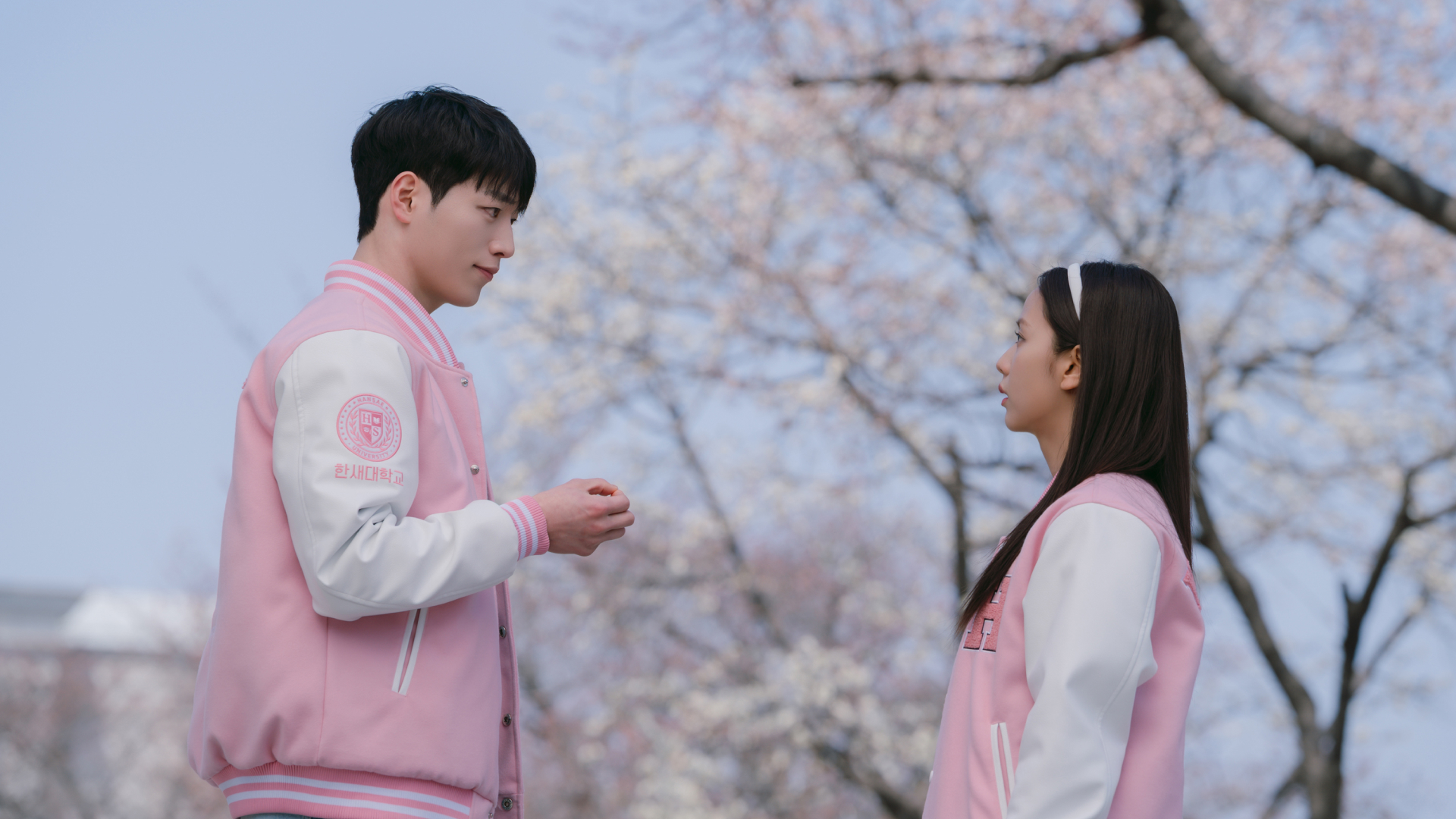 A man and a woman (Seo Kang-jun as Seo Eun-ho and Jisoo as Seo Mi-rae) wear pink and white Letterman jackets while standing outdoors in front of a cherry blossom tree, in 'Boyfriend on Demand.'