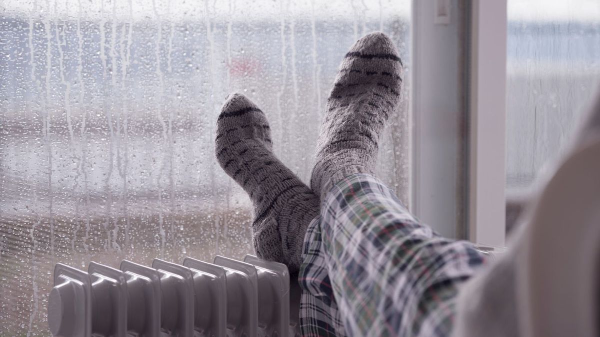 Woman wearing gray pair of woollen socks, warming cold feet in front of the heater, staying at home in the rainy winter season.