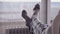 Woman wearing gray pair of woollen socks, warming cold feet in front of the heater, staying at home in the rainy winter season.
